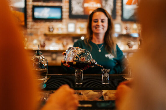 A smiling bartender pours a dark liquid from a large bottle into a shot glass at a bar, with bottles and decorations visible on shelves in the blurred background.