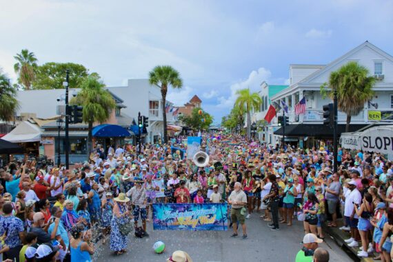 A large, colorful crowd fills a lively street during a festive parade, with palm trees, musicians, and banners under a partly cloudy sky, surrounded by vibrant shops and buildings.