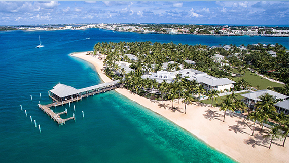 Aerial view of a tropical resort with white sandy beach, turquoise water, a wooden pier, and palm trees, with buildings nestled among greenery and a distant shoreline visible under a partly cloudy sky.
