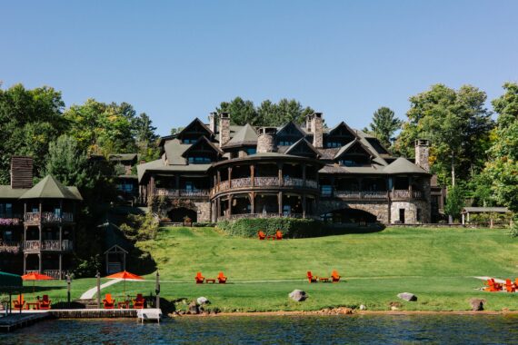 A large, rustic mansion with stone walls and multiple balconies sits on a grassy hill by a lake, surrounded by trees. Bright orange chairs are scattered on the lawn and at the water’s edge under umbrellas.