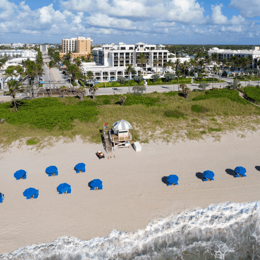 Aerial view of a sandy beach with rows of blue umbrellas near the shoreline, a lifeguard tower in the center, and hotels along a palm-lined street in the background under a partly cloudy sky.