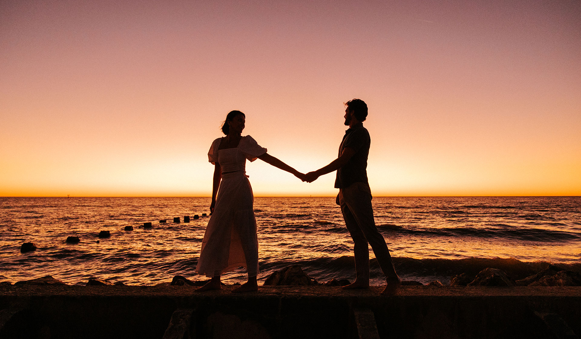 A couple holding hands stands on a rocky ledge by the ocean at sunset, silhouetted against the orange and purple sky—a perfect moment to experience what is shoulder season, with gentle waves in the background.