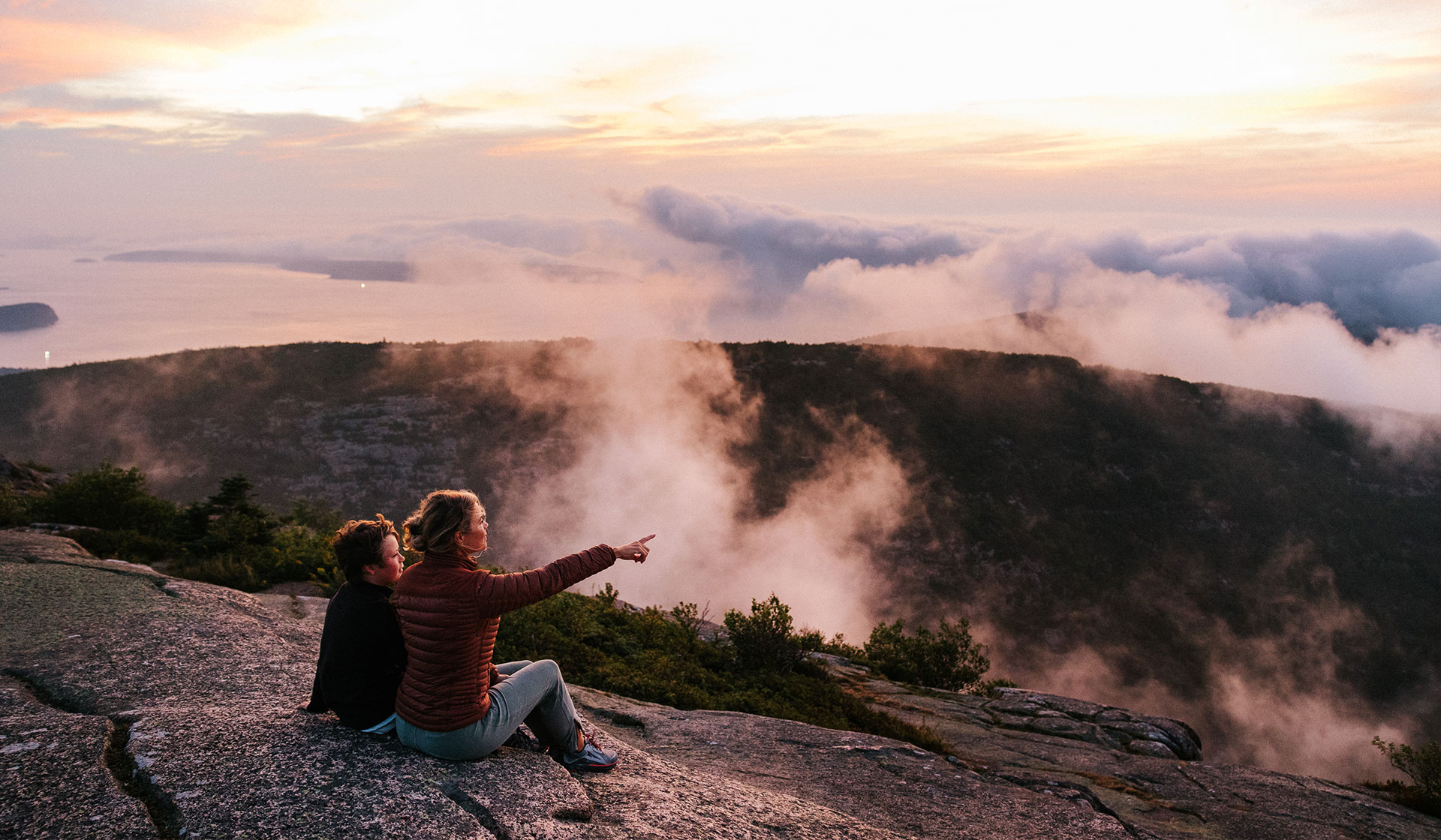 Two people sit on a rocky mountain ledge at sunrise or sunset during shoulder season. One points towards the horizon, where clouds drift over forested hills and water below, creating a peaceful and scenic atmosphere.