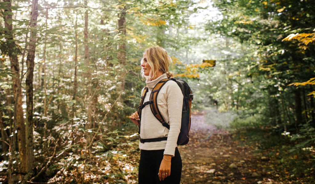 A person with blonde hair, wearing a light sweater and a backpack, stands on a sunlit forest path surrounded by green trees and foliage—an ideal moment to enjoy the peaceful beauty of shoulder season.