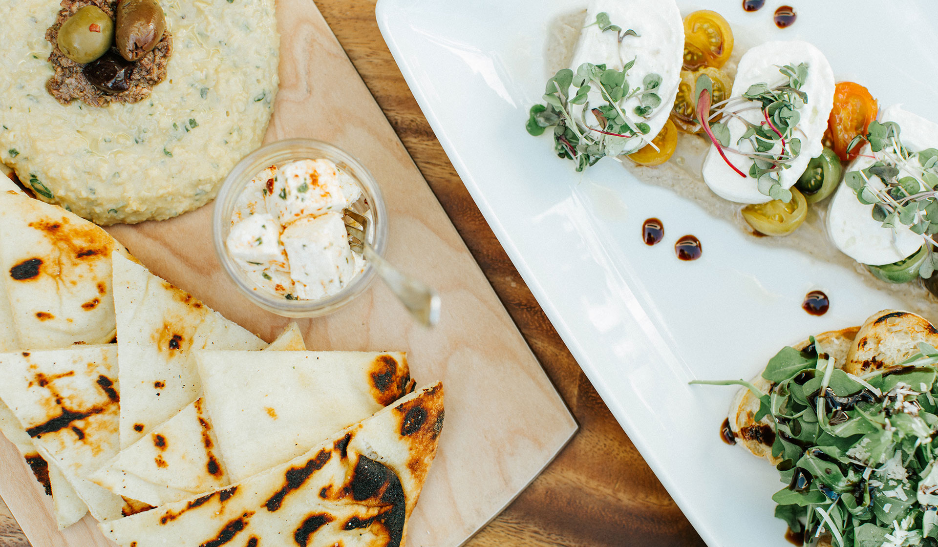 A platter with grilled flatbread, creamy dip with olives, a jar of marinated cheese, and a white plate of fresh mozzarella, cherry tomatoes, greens, and balsamic drizzle—perfect for savoring during the relaxing pace of shoulder season.