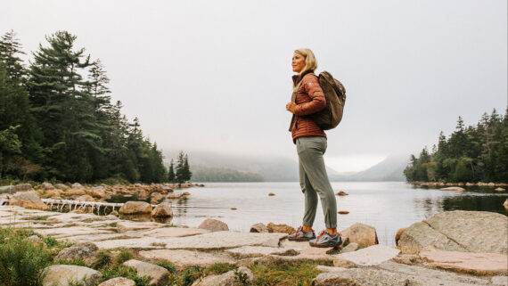 A woman wearing a backpack stands on a rocky shoreline by a calm lake, surrounded by pine trees and misty hills under an overcast sky, capturing the quiet beauty often found during shoulder season.