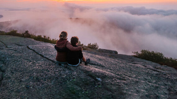 Two people sit close together on a rocky surface, overlooking a scenic landscape with clouds and a colorful sunset sky—a perfect moment to enjoy the peace of shoulder season, as shrubs and trees line the edge of the rocky area.