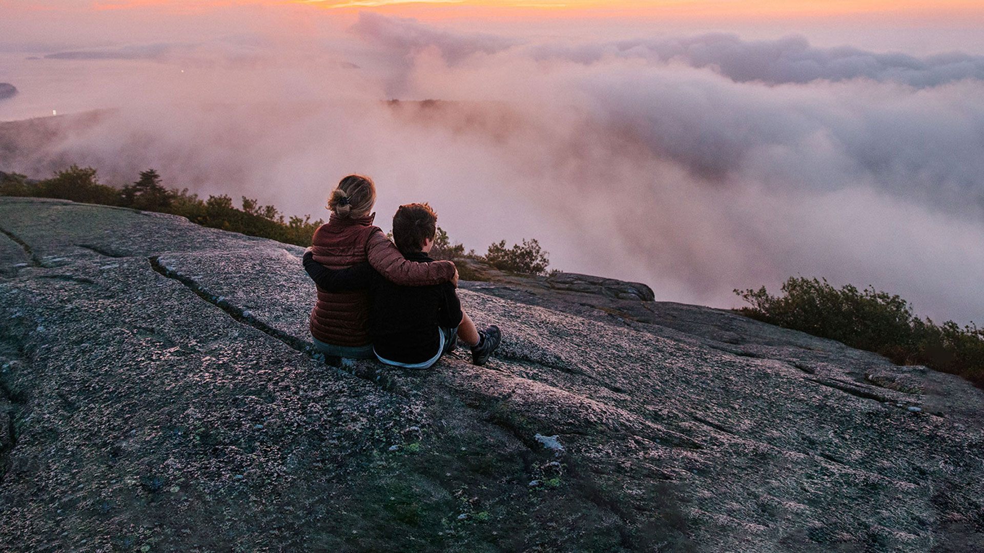 Two people sit close together on a rocky surface, overlooking a scenic landscape with clouds and a colorful sunset sky—a perfect moment to enjoy the peace of shoulder season, as shrubs and trees line the edge of the rocky area.