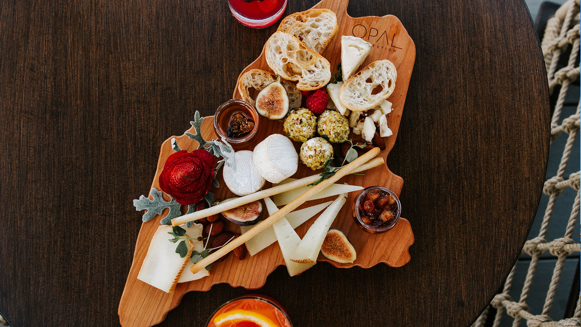 A wooden charcuterie board shaped like a state holds assorted cheeses, bread slices, figs, berries, breadsticks, nuts, leafy greens, and a small jar of jam, with colorful drinks on a round dark table nearby.