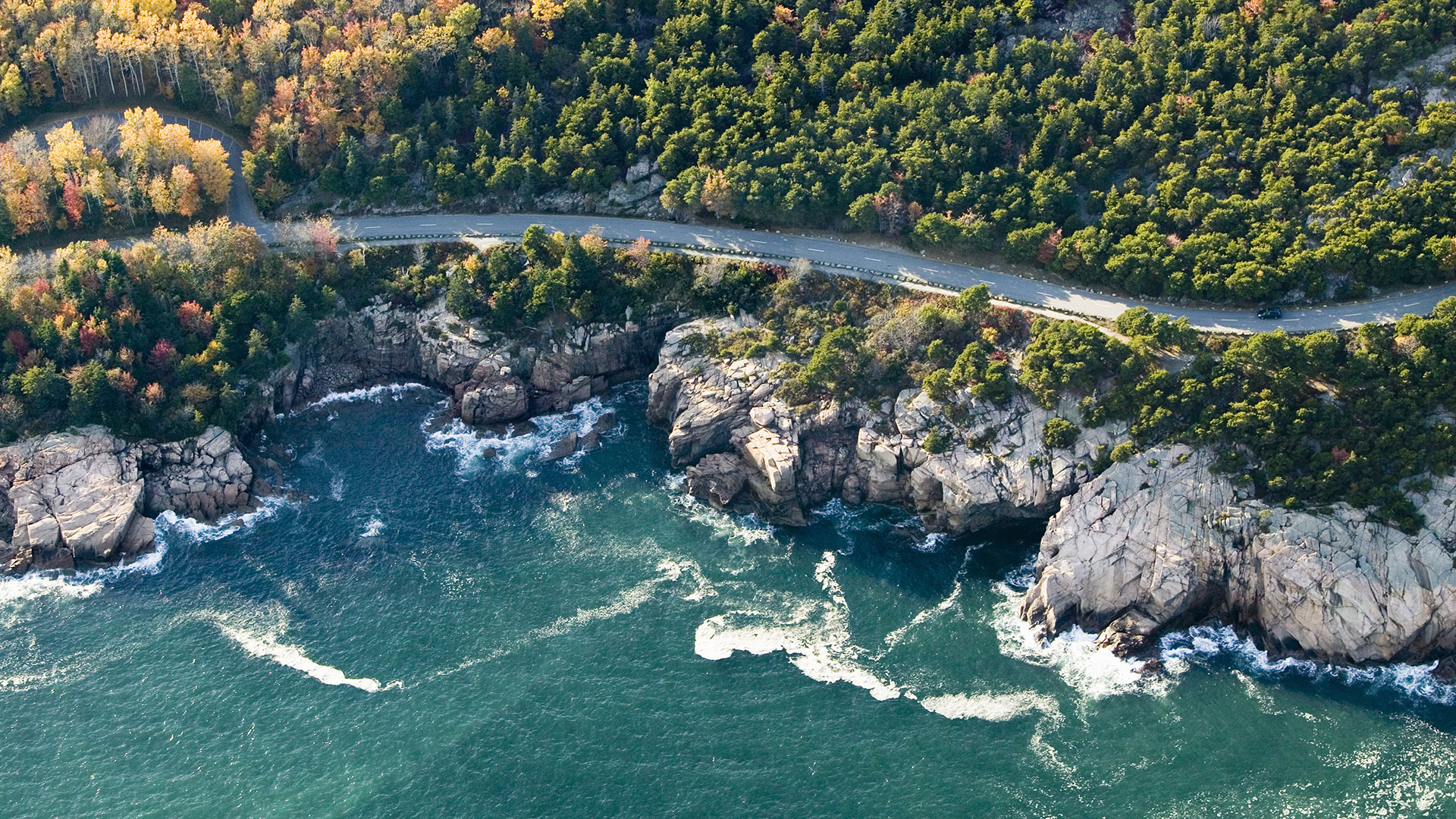 Aerial view of a winding coastal road bordered by dense forest with autumn foliage, rocky cliffs, and waves crashing against the shoreline below.