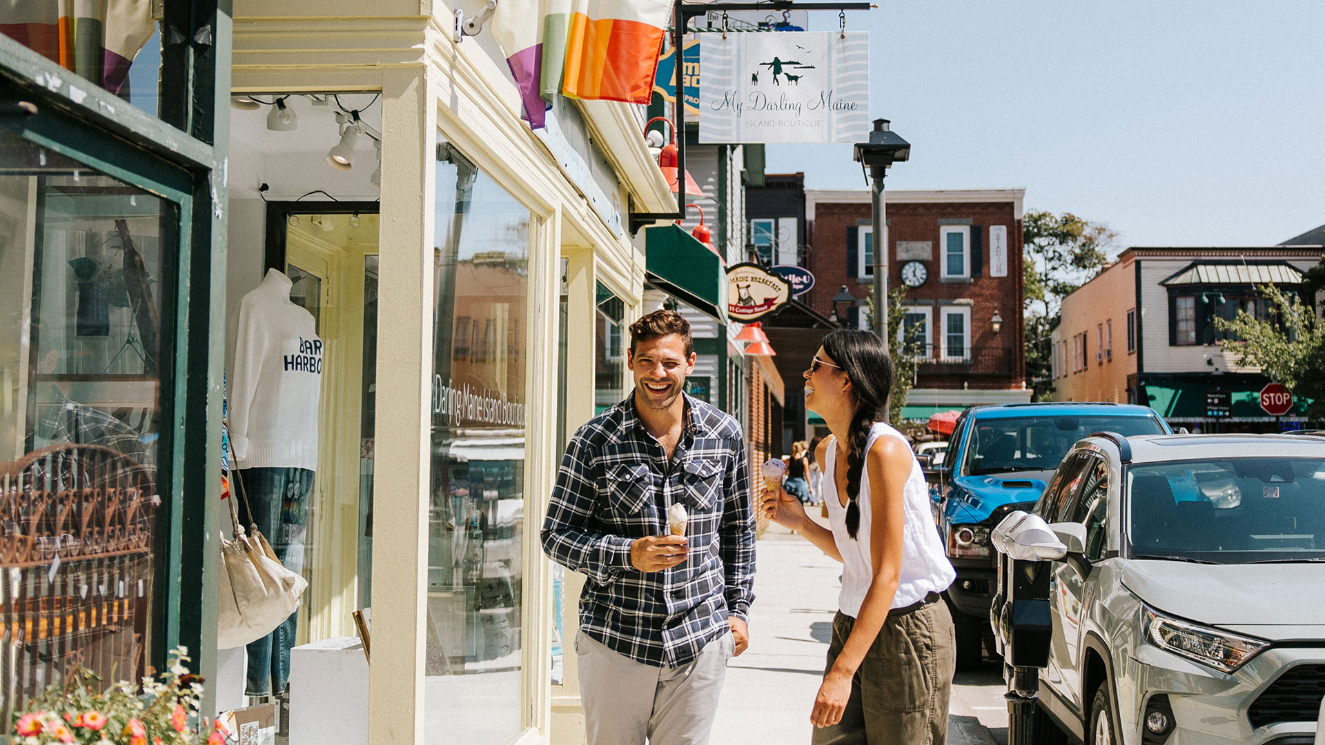 A man and woman smile and talk while walking down a sunny, lively street lined with shops, parked cars, and colorful flags. The man holds a drink, and they appear to be enjoying a leisurely day out.