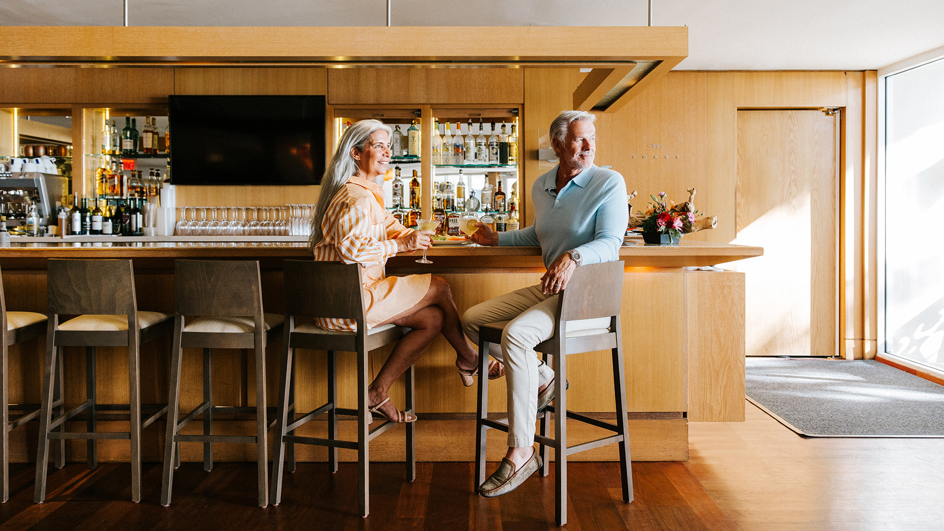 An older woman and man sit at the modern bar at Coast in Edgewater Beach Hotel with light wood décor, enjoying drinks together. The lively spot, like many Naples bars, is well-stocked and brightly lit.