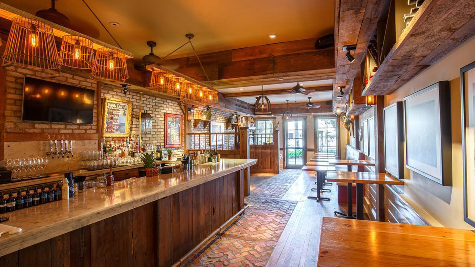 A warm, inviting Naples bar with exposed brick walls, a long wooden counter, hanging pendant lights, high tables, and large windows letting in natural light. The rustic decor and empty seating create the perfect setting for guests to unwind.