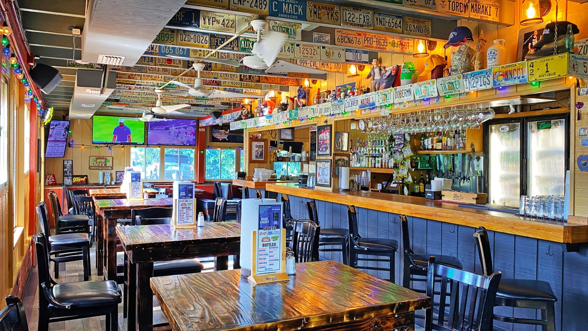 A brightly lit bar and restaurant with wooden tables and chairs, license plates lining the ceiling, TV screens on the walls, and bottles behind the counter. The empty space evokes the welcoming, casual vibe found in top Naples bars.