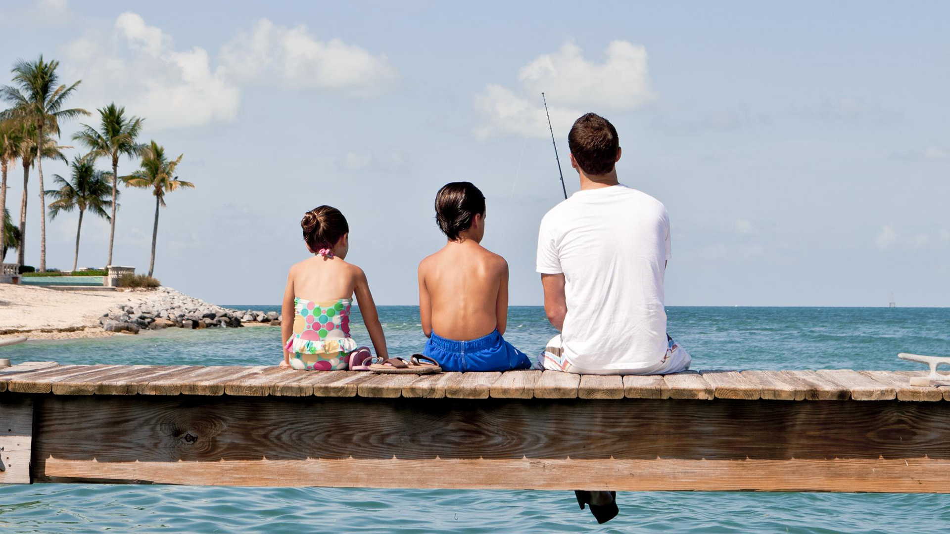 Three people, two children and one adult, sit on a wooden dock facing the ocean. The adult holds a fishing rod. Palm trees and a sandy beach are visible in the background under a partly cloudy sky.