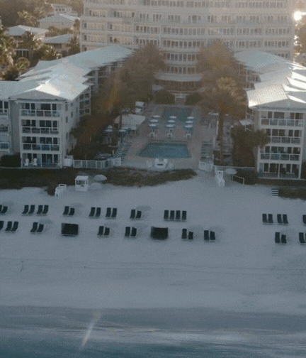 Aerial view of a beachfront resort with white buildings, a central pool area, lounge chairs arranged neatly on the sand, and gentle waves approaching the shore.
