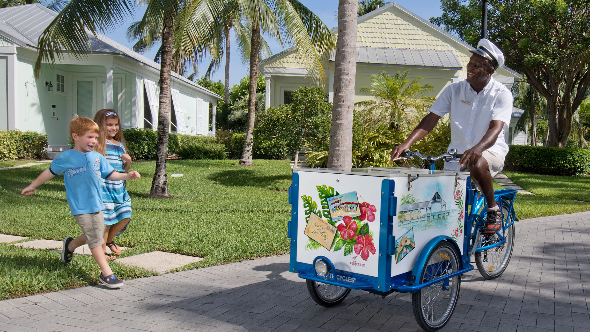 A man in a white shirt and hat rides an ice cream tricycle with a painted cooler in front, while two smiling children run toward him on a path lined with palm trees and pastel-colored houses—a perfect scene in Key West for kids.