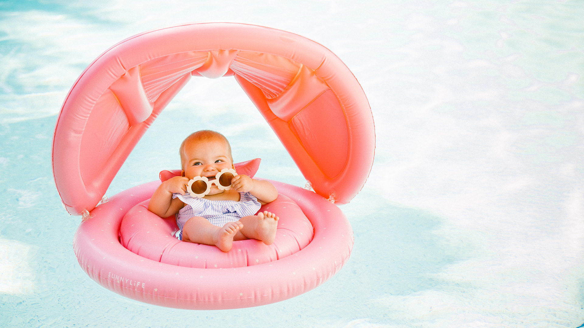 A baby wearing sunglasses sits in a pink inflatable float with a canopy, relaxing in a swimming pool under bright, clear light—perfect for a sunny key west for kids getaway.