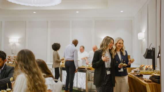 Business professionals gather around a buffet table in a bright, modern room, serving and enjoying food. Two women in front smile while holding plates, and chefs in the background prepare dishes.