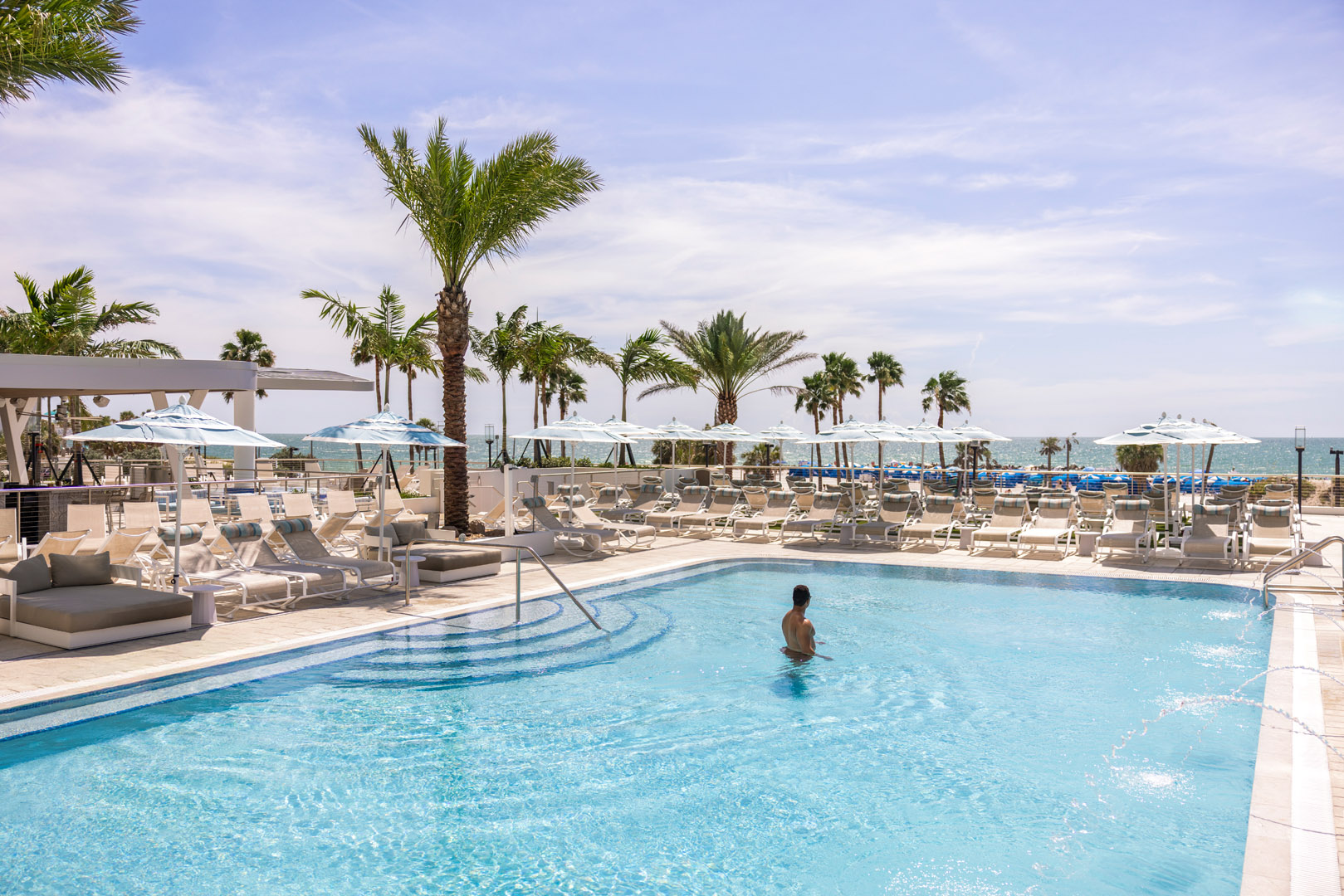 A person stands in a clear swimming pool surrounded by lounge chairs, umbrellas, and palm trees, with the ocean and blue sky visible in the background.