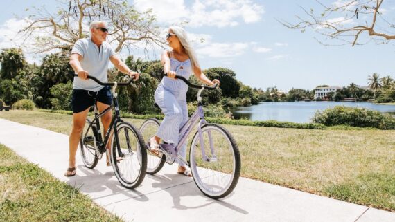 An older man and woman ride bicycles side by side on a sunny path near a lake, smiling at each other. Trees, grass, and waterfront homes are visible in the background—enjoying one of the many relaxing things to do in Naples under a blue sky.