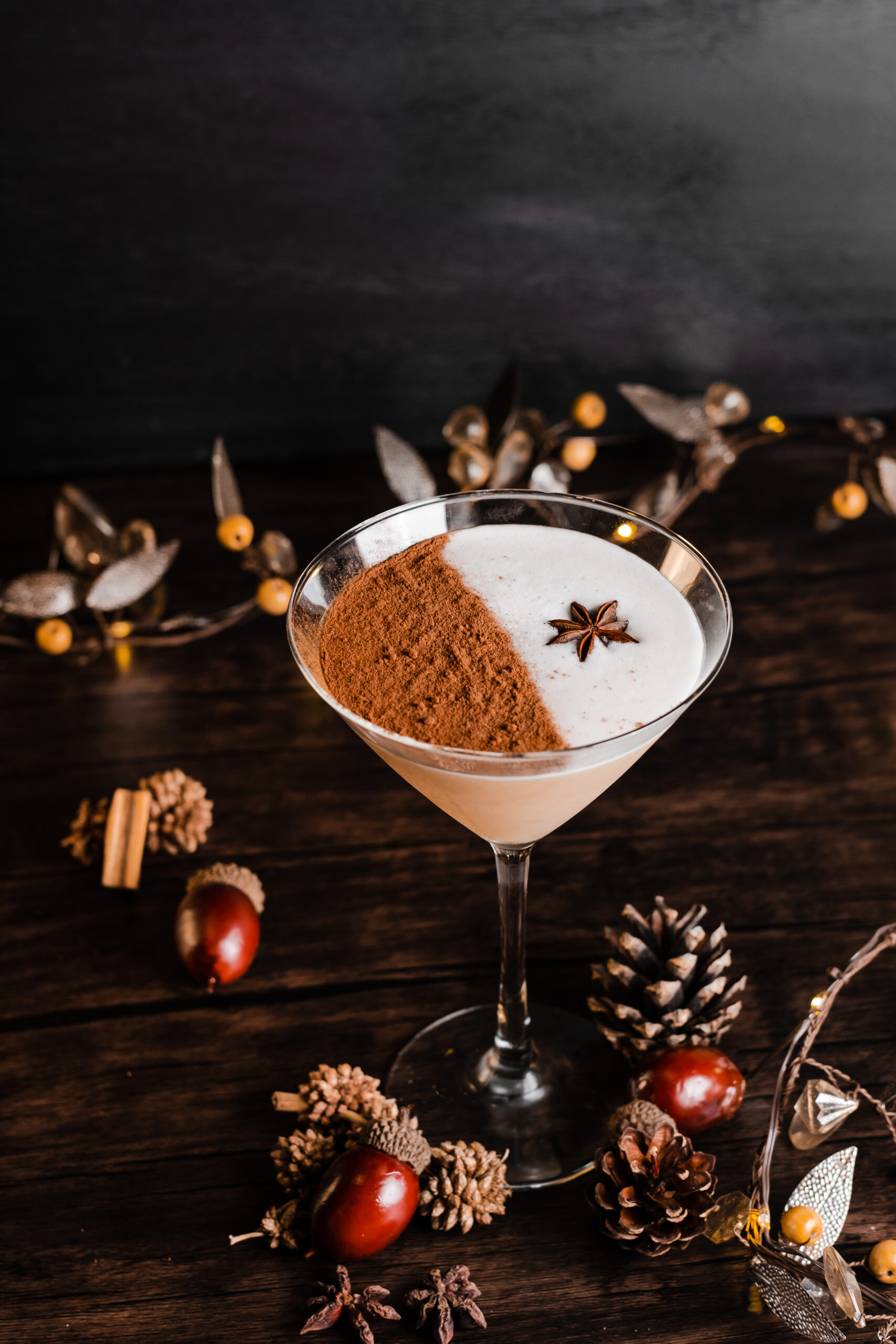 A festive cocktail in a martini glass is garnished with a dusting of cinnamon on one half and a star anise on top. The wooden table is decorated with pinecones, acorns, cinnamon sticks, and holiday ornaments.