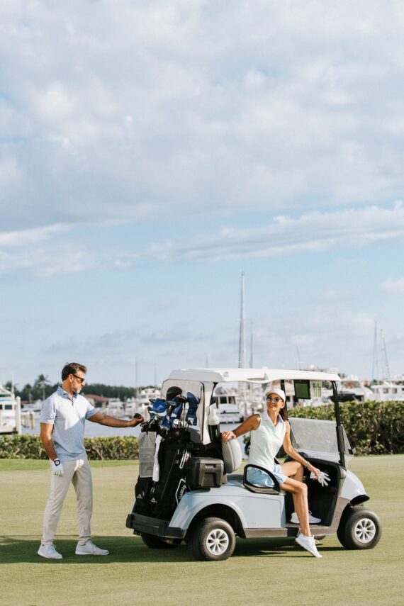A man stands beside a golf cart while a woman sits in the driver’s seat, both dressed in golf attire on a sunny day, with a marina and boats in the background.