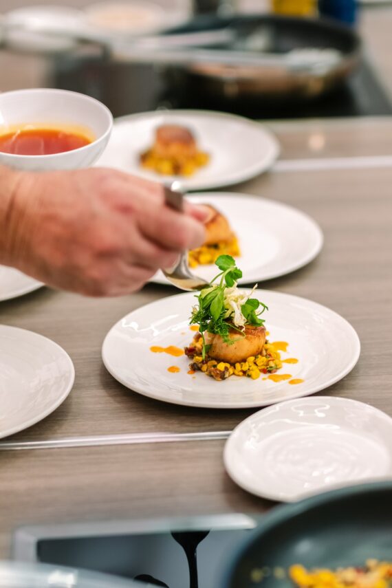 A chef’s hand adds garnish to a plated dish of seared scallop on corn and vegetable medley, with vibrant sauce, on a white plate in a professional kitchen setting.