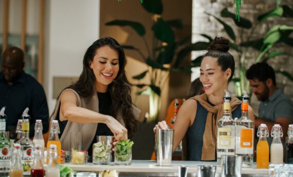 Two women stand at a bar counter, smiling while preparing drinks with fresh ingredients. Bottles of alcohol and mixers are on the counter. Other people are blurred in the background, and plants decorate the space.