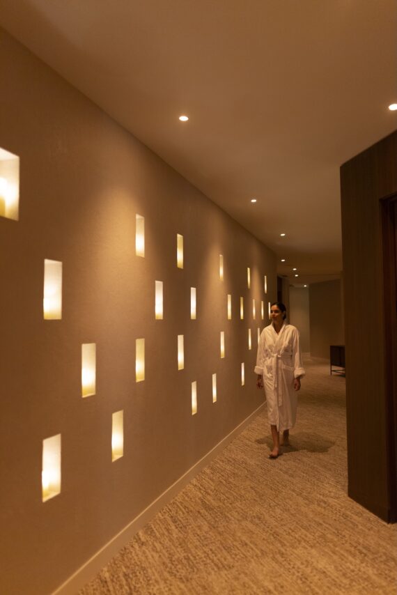 A person in a white robe walks barefoot down a softly lit hallway with rectangular wall sconces emitting warm light. The atmosphere is calm and relaxing, suggesting a spa or wellness center.