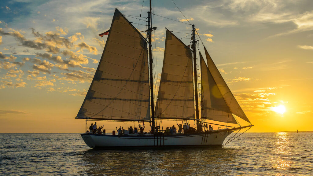 A large sailboat with two tall sails glides on the water at sunset. The sky is golden, and people on the boat are waving as the sun sets on the horizon, casting a warm glow over the ocean.