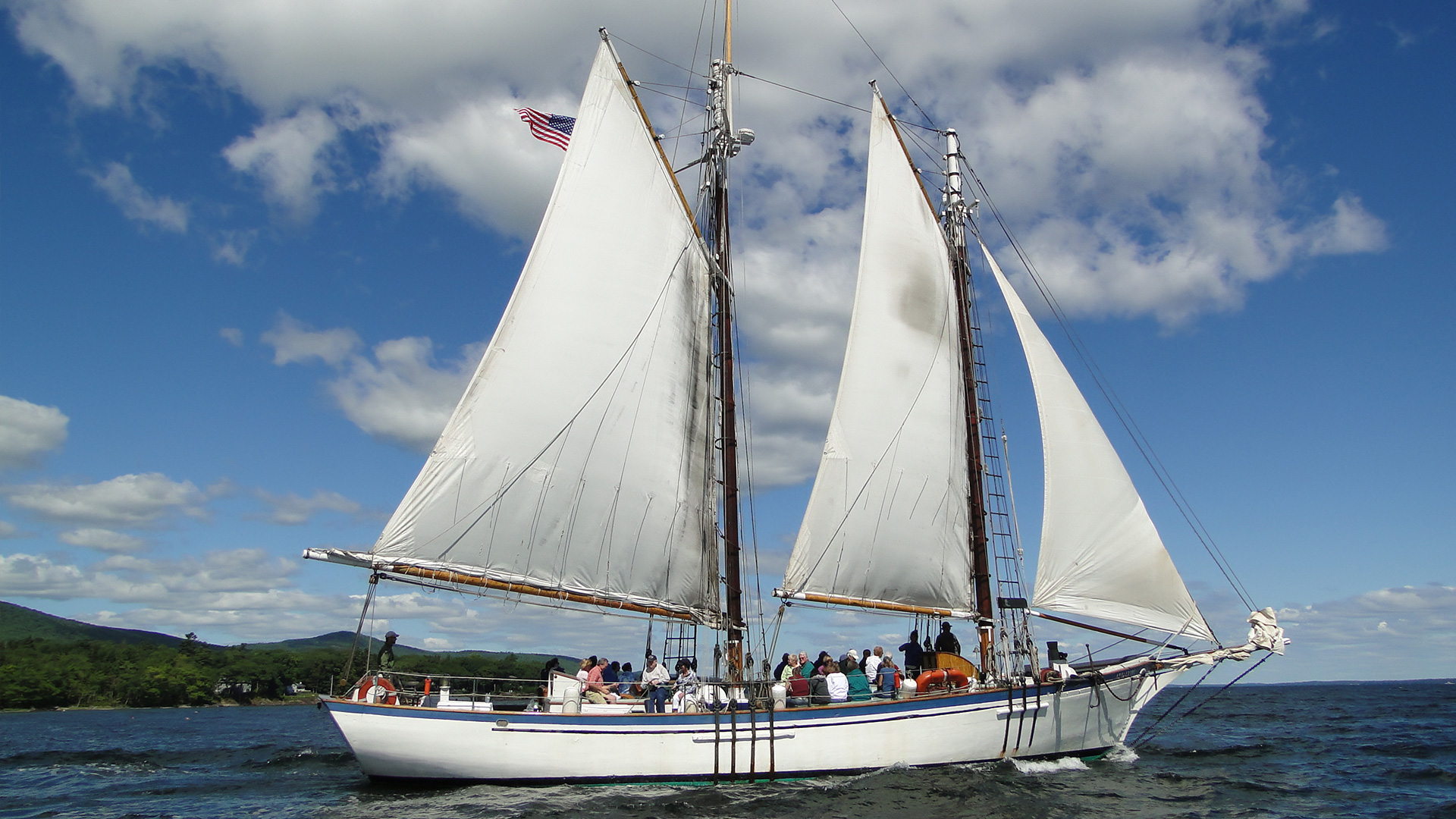 A white sailboat with two tall masts and large sails carries a group of people on a sunny day, sailing on a blue lake with green hills and a partly cloudy sky in the background. An American flag flies at the stern.