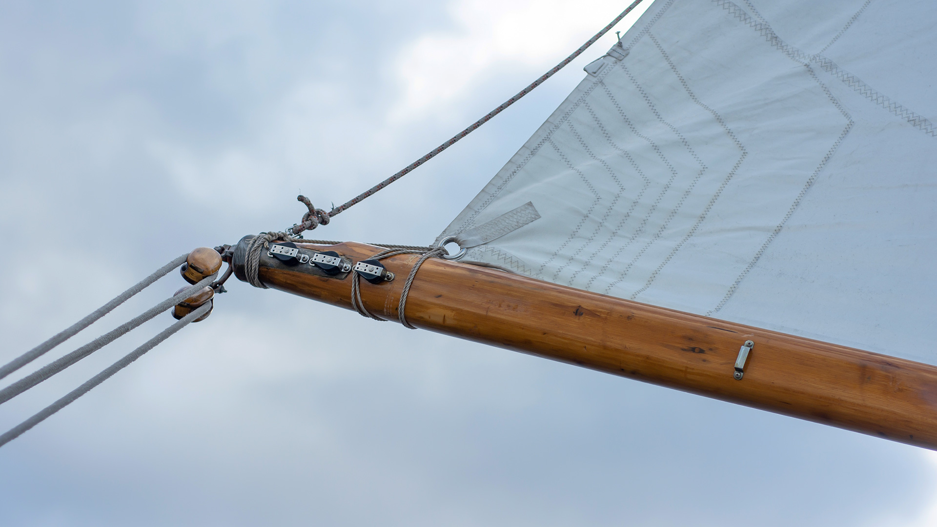Close-up of a sailboat’s wooden boom and white sail against a cloudy sky, showing ropes, pulleys, and metal fittings attached to the sail and boom.