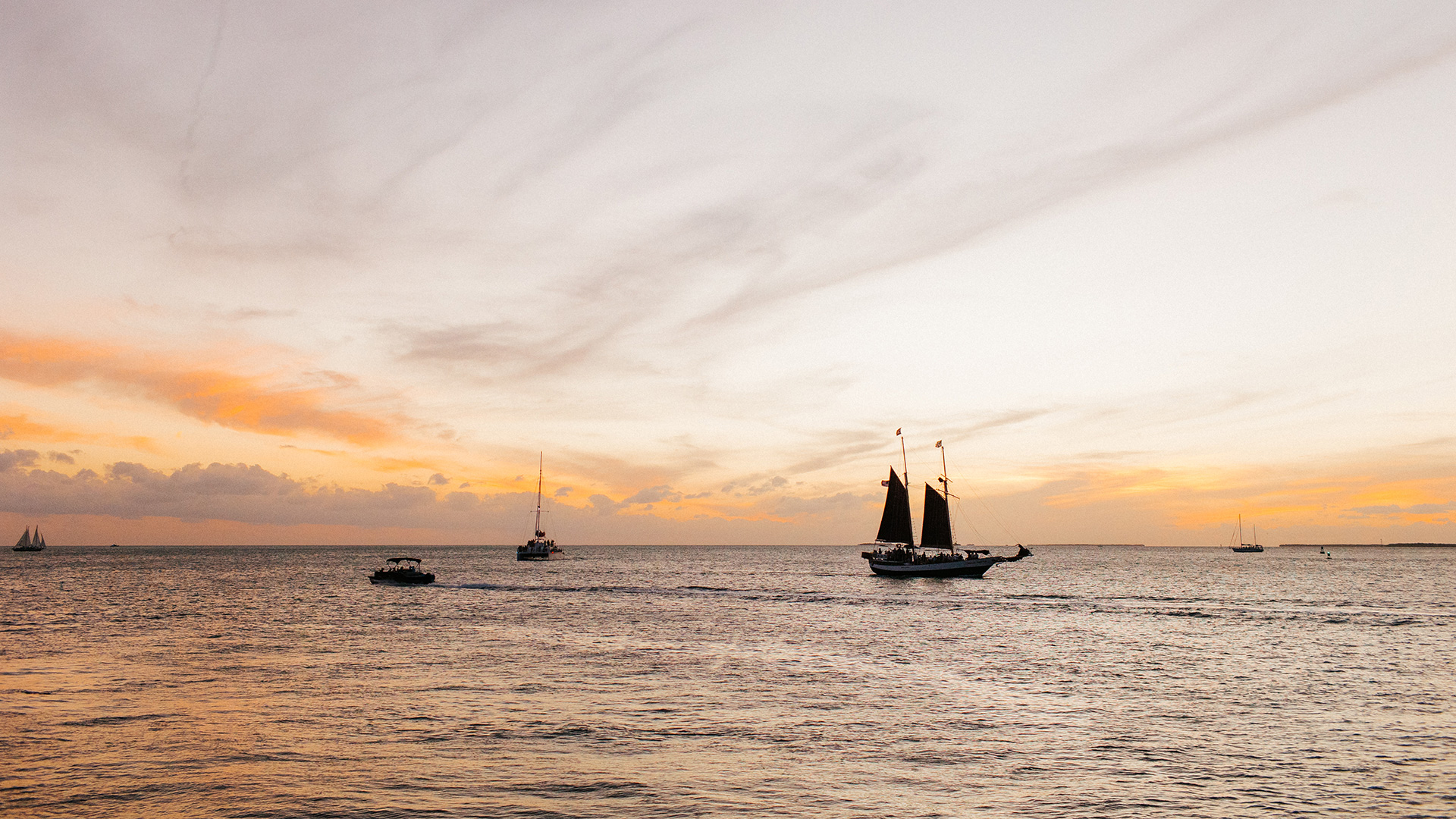 Sailboats and small boats float on calm water at sunset, with soft orange and pink hues in the cloudy sky. The scene is tranquil and serene, with boats silhouetted against the horizon.