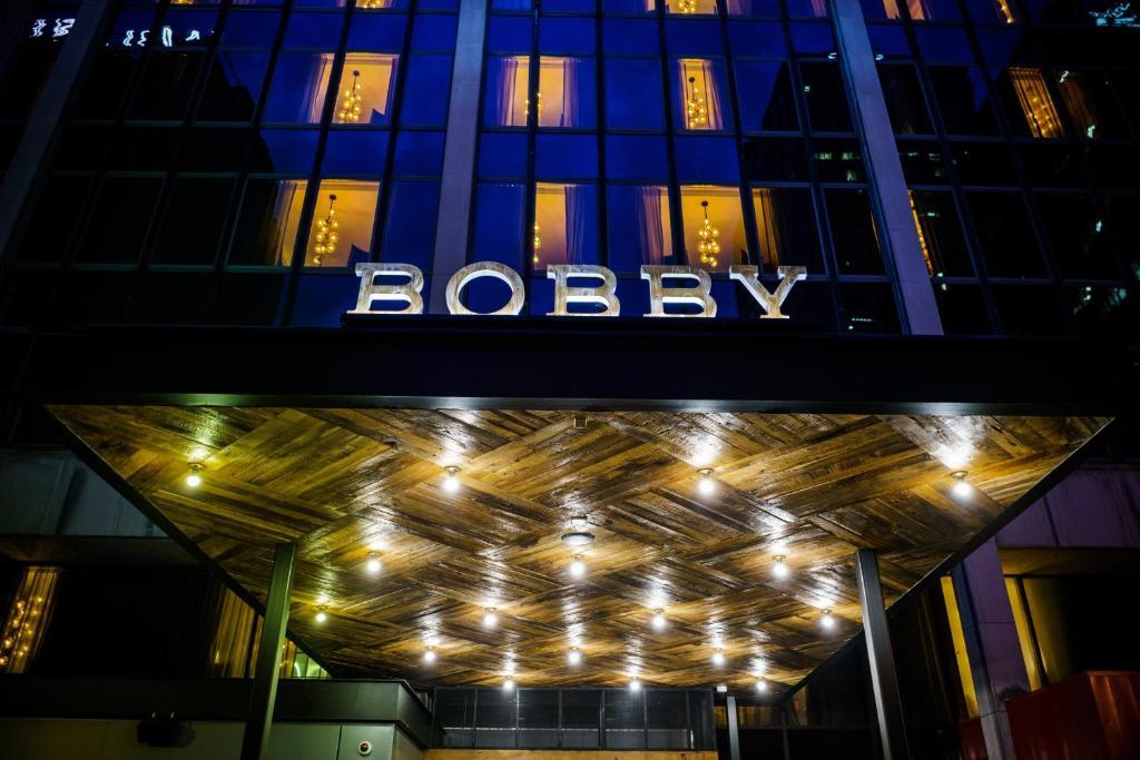 The entrance of a modern building with a wooden ceiling and lights, featuring large illuminated letters spelling BOBBY above the doorway, with tall glass windows reflecting lights at night.
