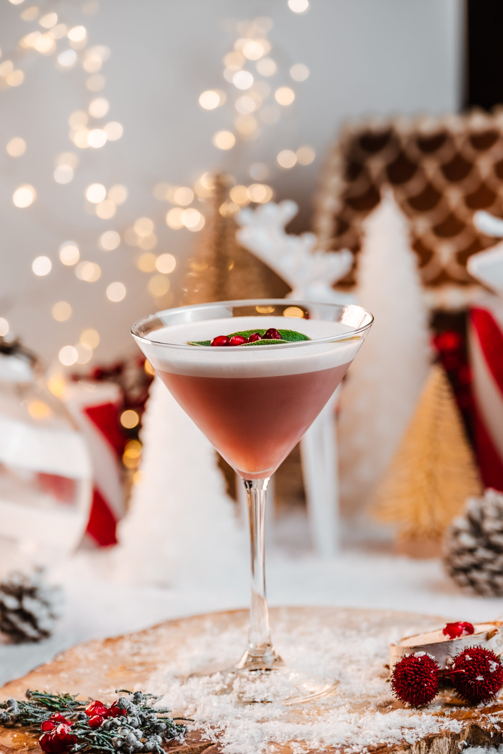 A festive pink cocktail in a martini glass, garnished with mint leaves and red berries, sits on a snowy surface. Blurred holiday decorations, pinecones, and twinkling lights create a cozy, wintery background.