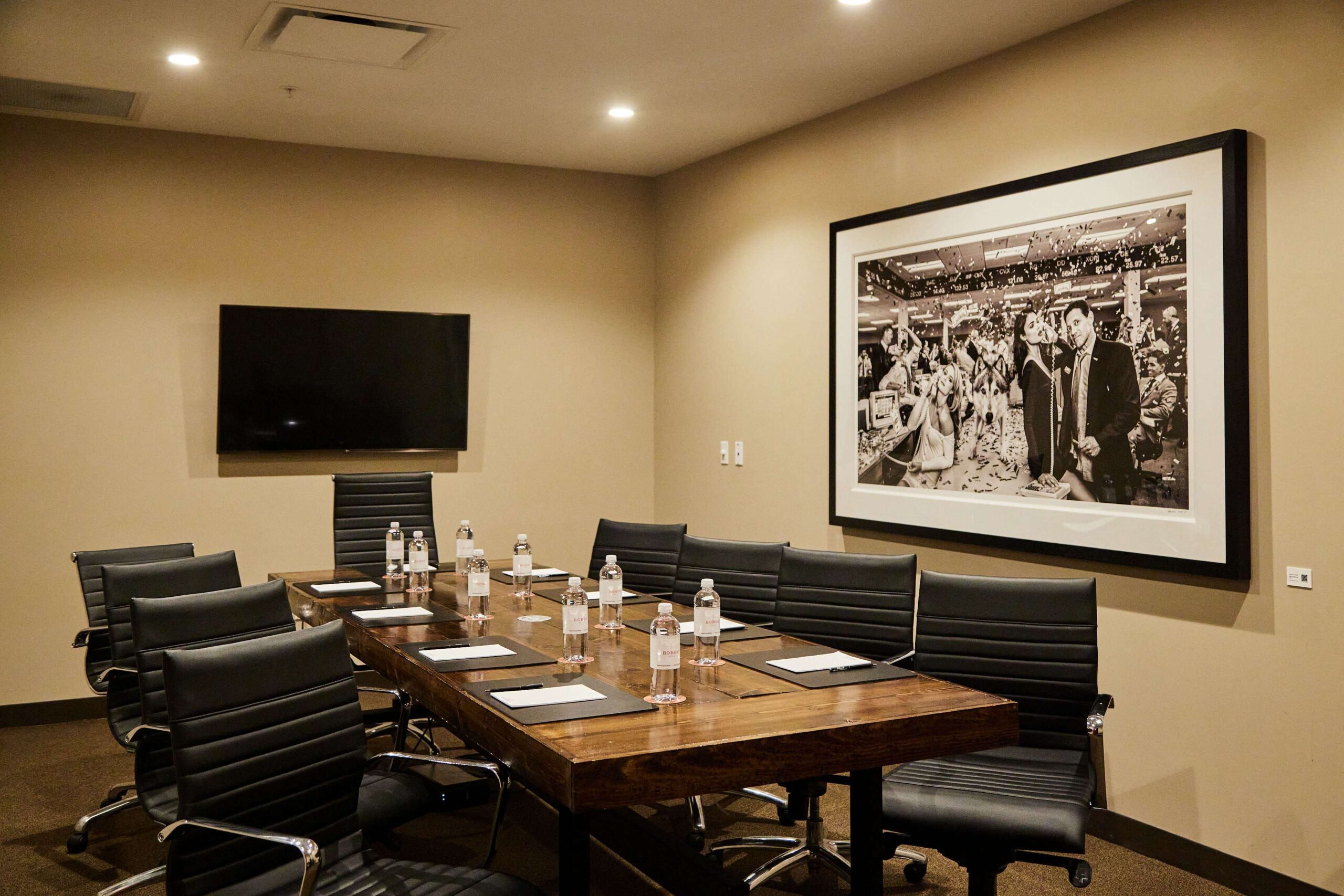 A conference room with a wooden table, black office chairs, bottled water, notepads, a wall-mounted TV, and a large black-and-white photo on the beige wall.