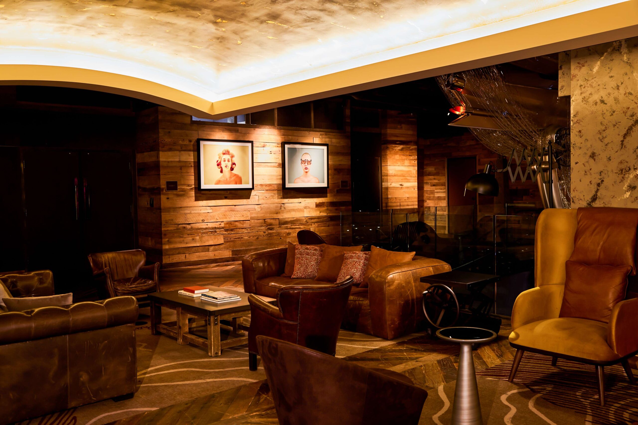 A cozy lounge with leather chairs and sofas, wooden walls, warm lighting, and framed portraits on the wall, creating an inviting and stylish atmosphere.