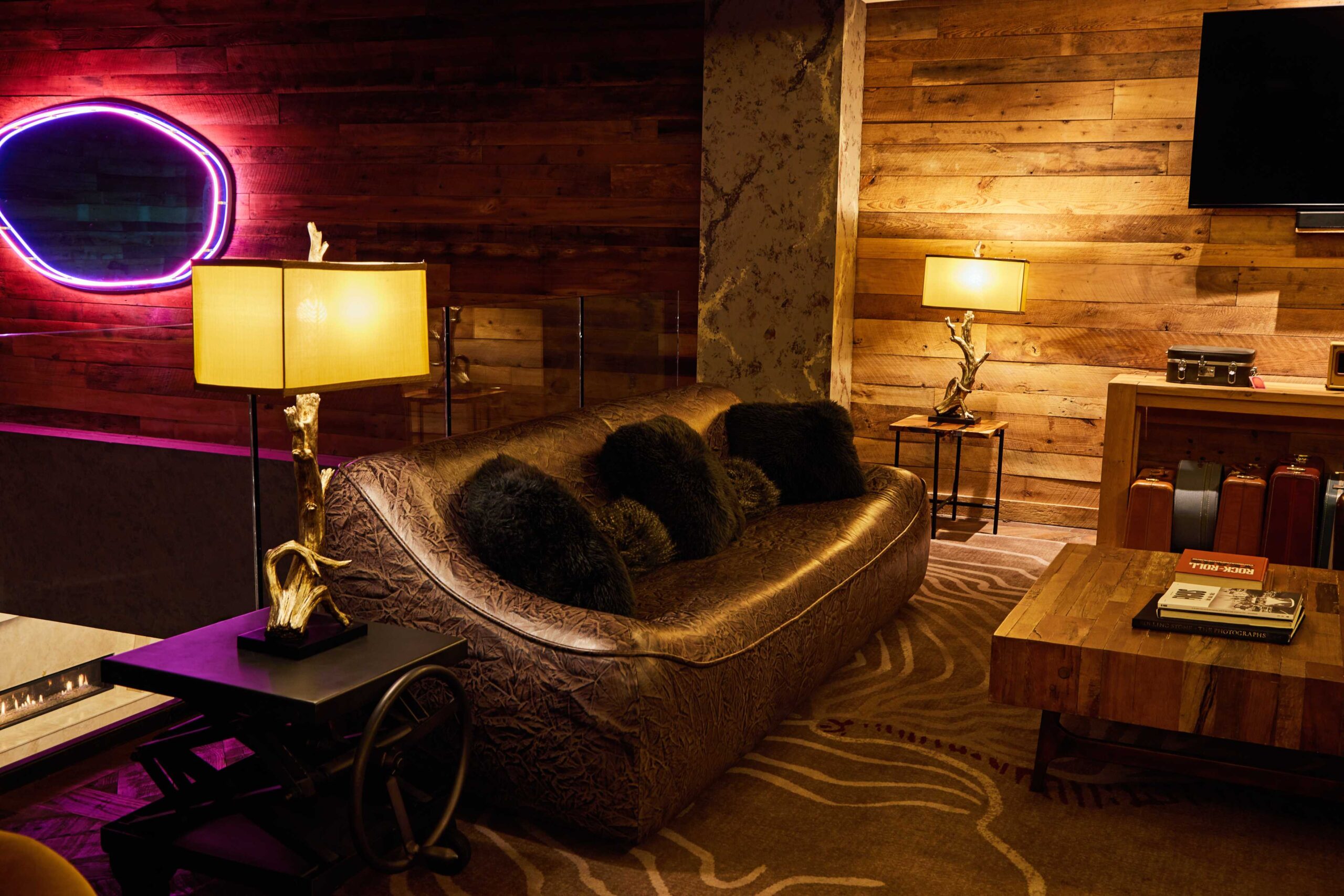 A cozy living room with a brown textured sofa, black fur pillows, two unique lamps with gold bases, a rustic wooden coffee table, and wood-paneled walls. A neon light and TV hang on the wall.