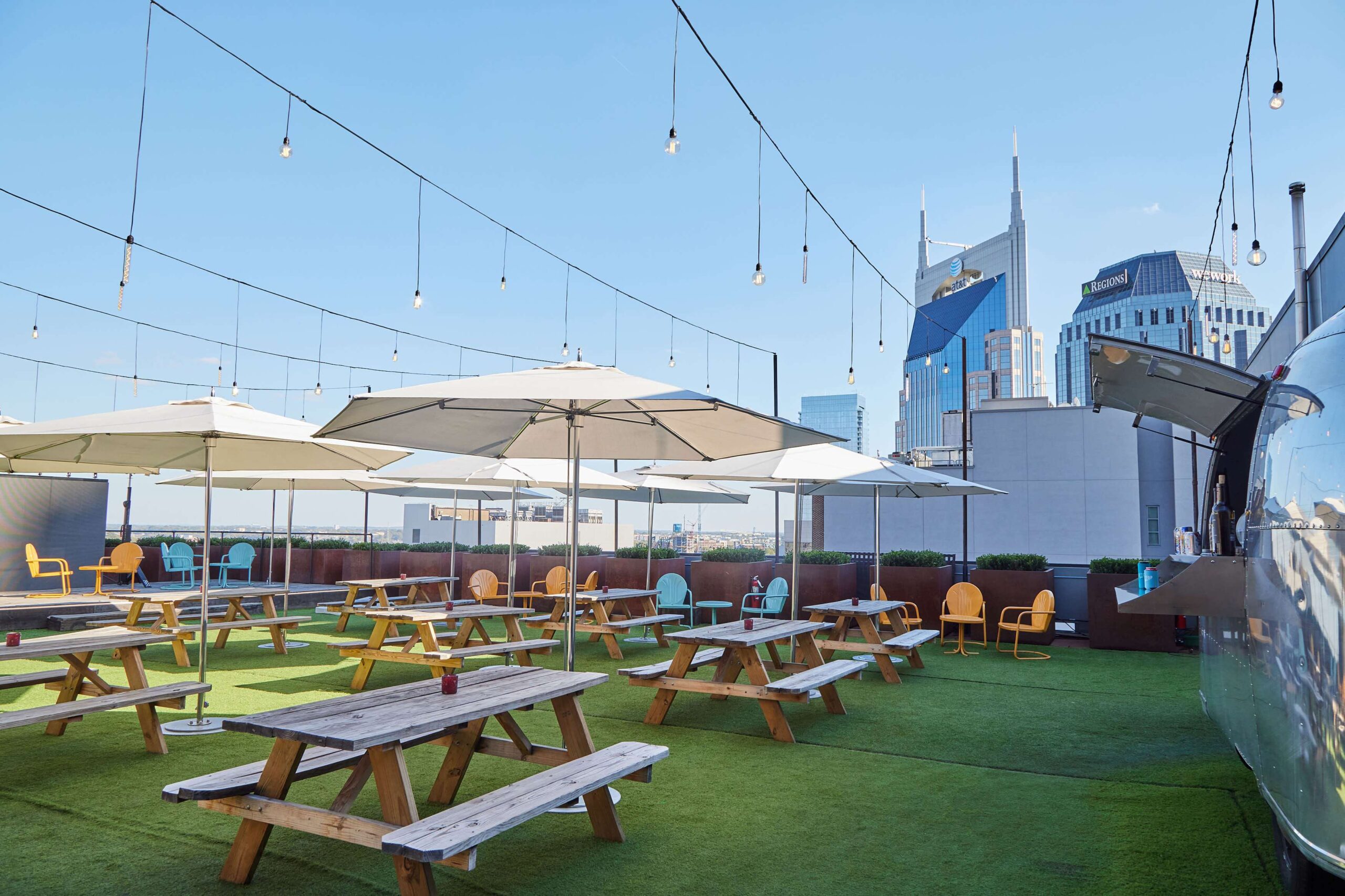 Rooftop patio with picnic tables, umbrellas, and string lights, set on green artificial grass. City skyscrapers are visible in the background under a clear blue sky.