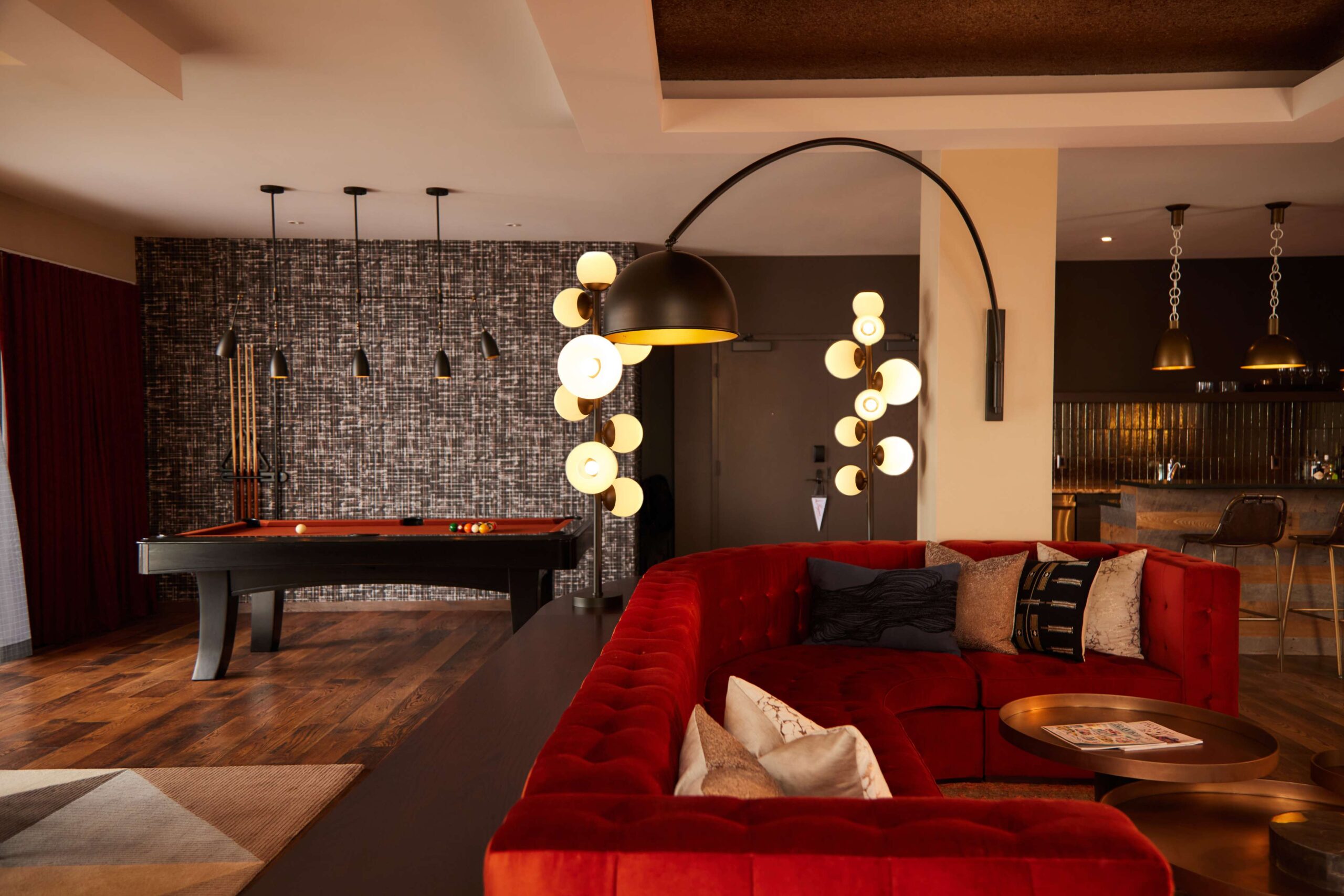 A stylish lounge area with a red velvet sectional sofa, a modern floor lamp with multiple globes, a pool table, and a kitchen with bar stools in the background. Warm lighting creates a cozy atmosphere.