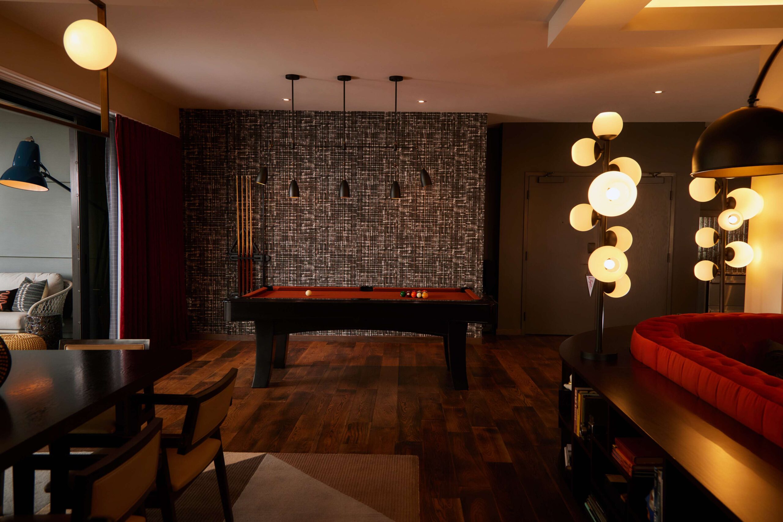 A stylish game room with a red pool table, hanging pendant lights, wooden floor, cue sticks on the wall, modern lamps, and a cozy seating area partially visible on the left.