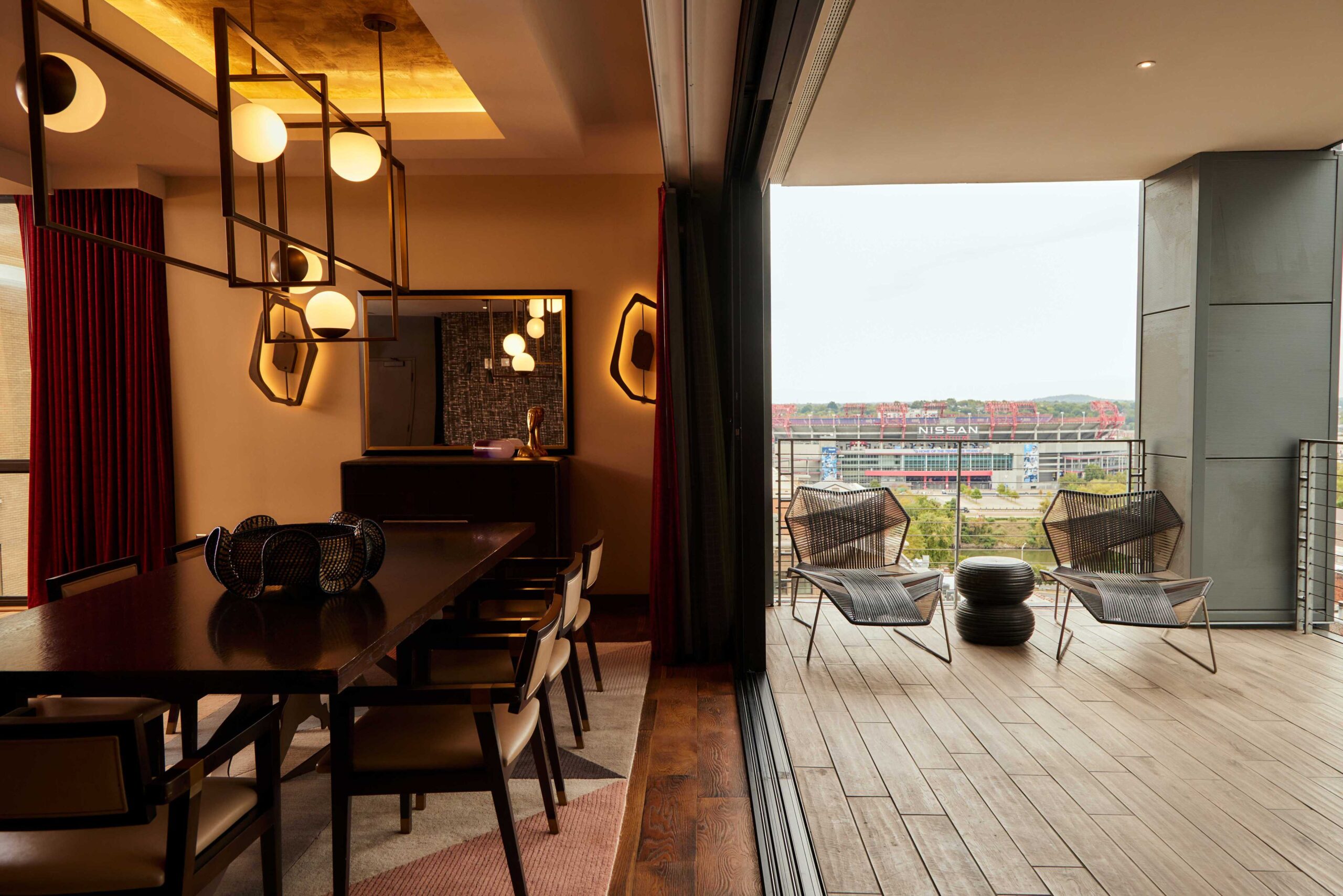 Modern dining area with geometric light fixtures and a dark wood table, opening onto a spacious balcony with two lounge chairs and a view of a stadium in the distance.