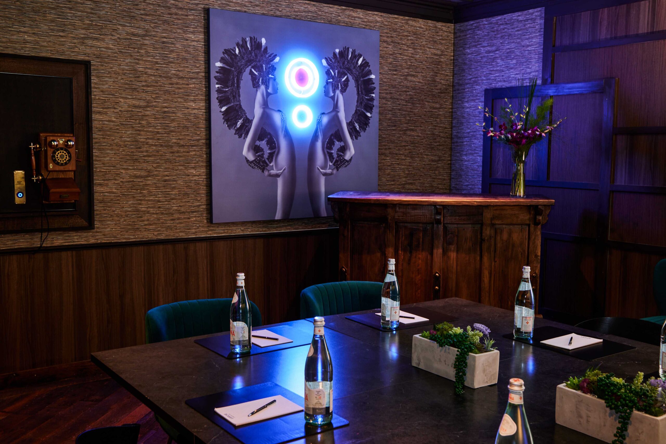 A stylish meeting room with a large table set with bottled water, notepads, and pens. On the wall is modern art of mirrored figures with neon lights, and a vintage phone is mounted nearby. A floral arrangement is on a wooden cabinet.