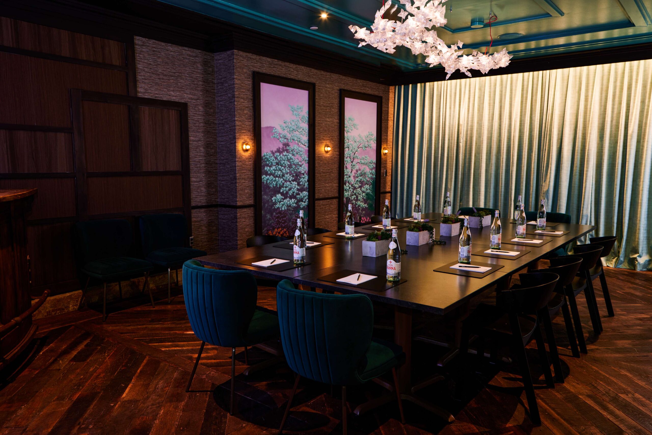 A dimly lit conference room with a long table set for a meeting, featuring green velvet chairs, water bottles, notepads, pens, decorative wall panels with tree art, and a modern white chandelier.