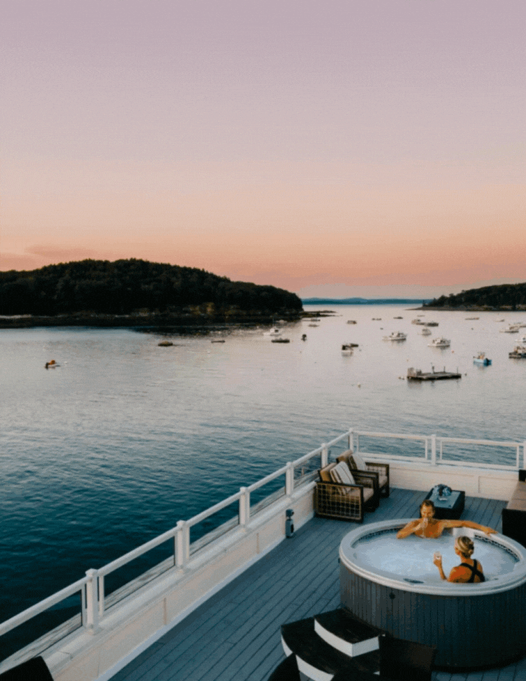 Two people relax in a hot tub on a deck overlooking a calm bay with boats, surrounded by lounge chairs, at sunset with a pink and purple sky above distant tree-covered hills.