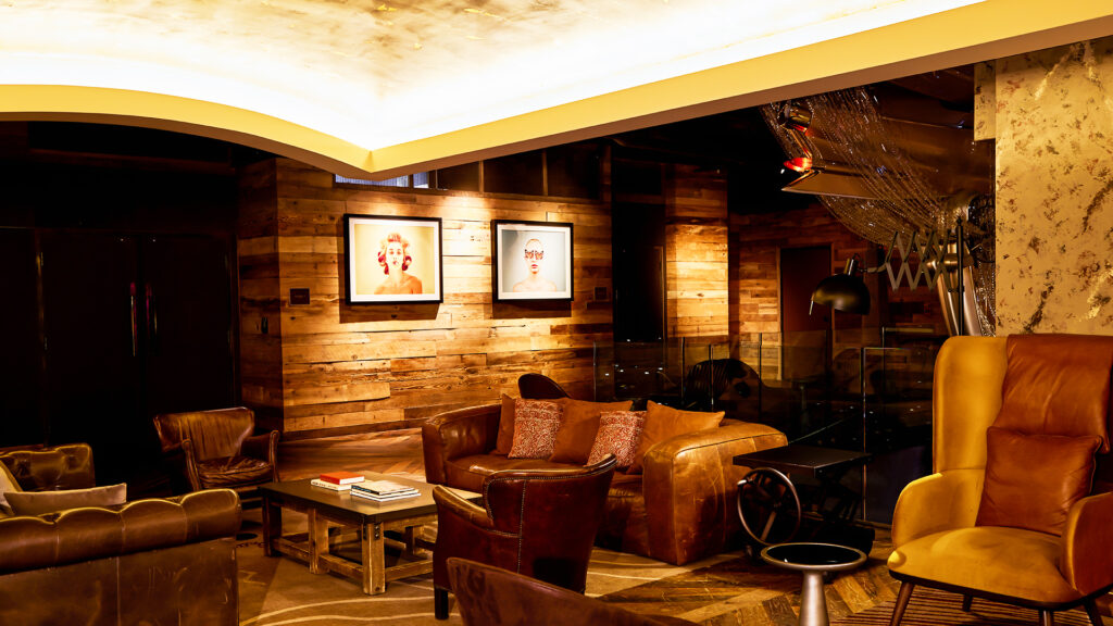 A cozy lounge with warm lighting, featuring leather armchairs, wooden walls, framed portraits, a coffee table with books, and modern decor elements.