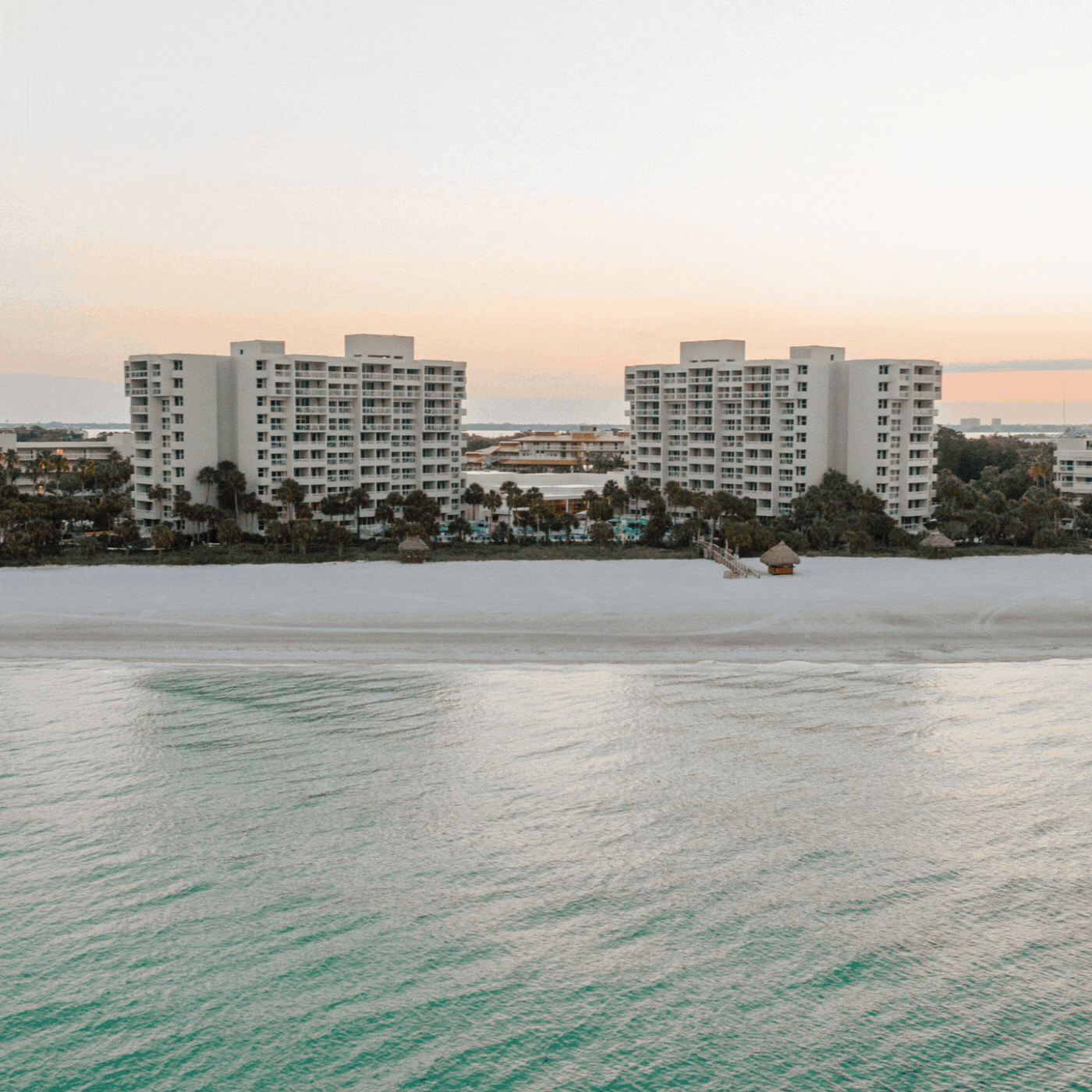A wide sandy beach bordered by calm turquoise water, with two large white condominium buildings and palm trees in the background under a pale sky at sunset.