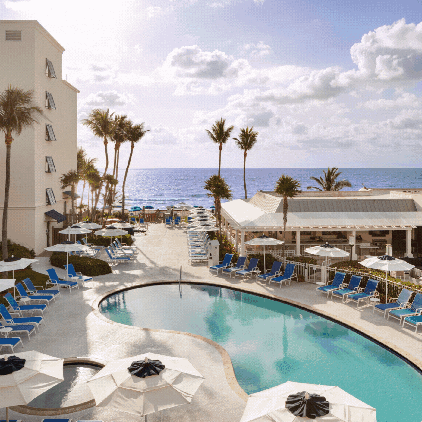 A beachside resort featuring a curvy pool surrounded by blue lounge chairs and white umbrellas, palm trees, and a view of the ocean under a partly cloudy sky.