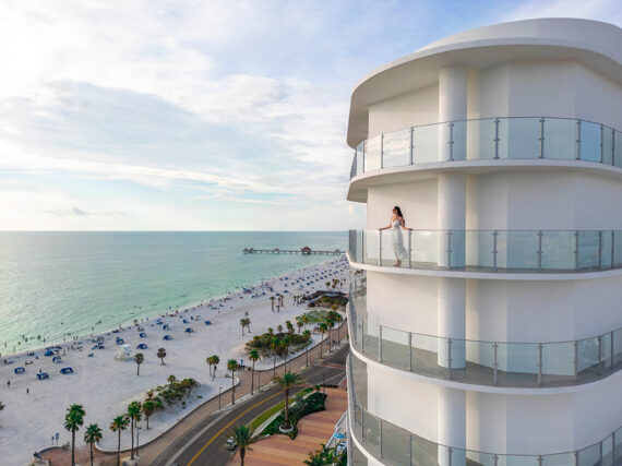 A person stands on a curved balcony of a modern white building overlooking a sandy beach, palm trees, and calm ocean waters under a partly cloudy sky.